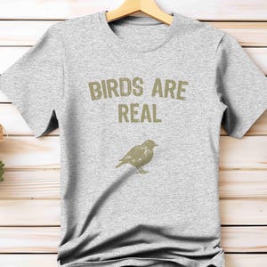 K&ouml;nnte beinhalten: Hellgraues T-Shirt mit dem Text "BIRDS ARE REAL" in hellbrauner Farbe und einer Vogelillustration. Das T-Shirt h&auml;ngt an einem Holzb&uuml;gel vor einem hellen Holz Hintergrund.