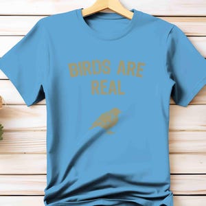 K&ouml;nnte beinhalten: Hellblaues T-Shirt mit dem goldenen Schriftzug "BIRDS ARE REAL" und einer goldenen Vogelgrafik. Das T-Shirt wird auf einem Holzkleiderb&uuml;gel pr&auml;sentiert.