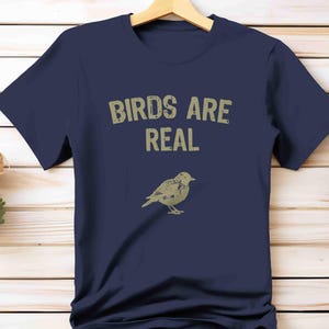 K&ouml;nnte beinhalten: Marineblaues T-Shirt mit dem Text "BIRDS ARE REAL" in verblichener, hellbrauner Schrift. Darunter eine detaillierte Illustration eines Vogels. Das T-Shirt h&auml;ngt an einem Holzb&uuml;gel.
