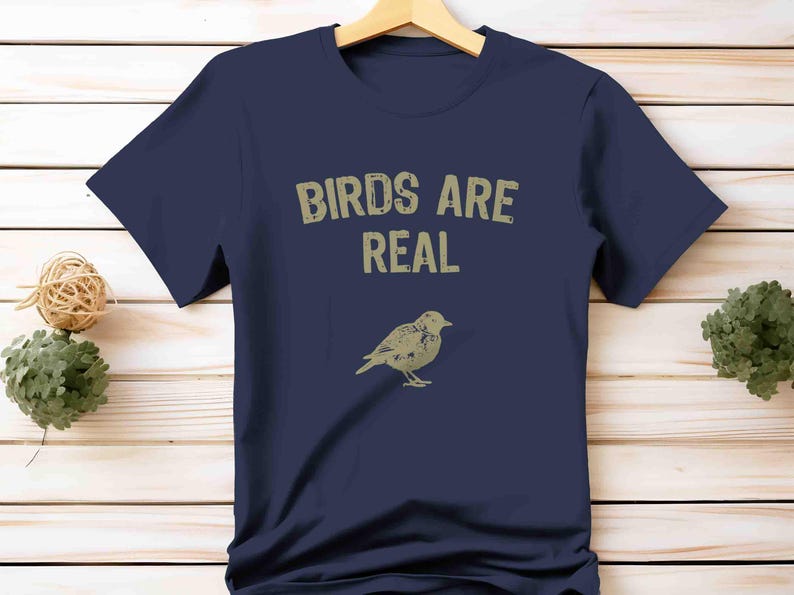 K&ouml;nnte beinhalten: Marineblaues T-Shirt mit dem Text "BIRDS ARE REAL" in verblichener, hellbrauner Schrift. Darunter eine detaillierte Illustration eines Vogels. Das T-Shirt h&auml;ngt an einem Holzb&uuml;gel.