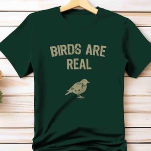 K&ouml;nnte beinhalten: Dunkelgr&uuml;nes T-Shirt mit dem Text "BIRDS ARE REAL" in verblichener Schrift &uuml;ber einer beigen Vogelillustration. Das T-Shirt h&auml;ngt an einem Holzkleiderb&uuml;gel.