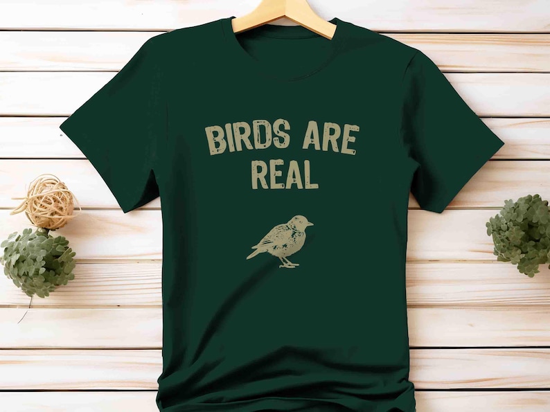 K&ouml;nnte beinhalten: Dunkelgr&uuml;nes T-Shirt mit dem Text "BIRDS ARE REAL" in verblichener Schrift &uuml;ber einer beigen Vogelillustration. Das T-Shirt h&auml;ngt an einem Holzkleiderb&uuml;gel.
