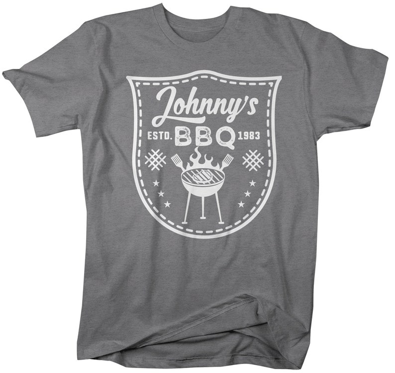 May include: A gray t-shirt with a white graphic featuring the text "Johnny's BBQ Estd. 1983". The graphic includes a shield shape, a grill, flames, and stars, perfect for barbecue enthusiasts.