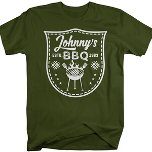 May include: Olive green t-shirt featuring a white graphic. The graphic includes the text "Johnny's BBQ ESTD. 1983" inside a shield design, along with a grill and stars. A casual tee for barbecue lovers.