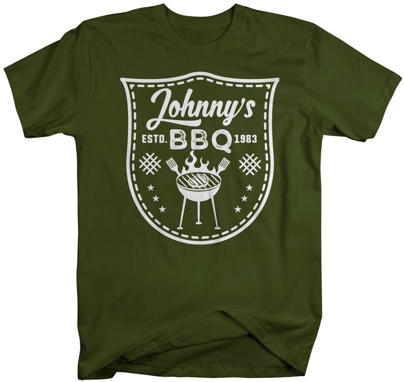 May include: Olive green t-shirt featuring a white graphic. The graphic includes the text "Johnny's BBQ ESTD. 1983" inside a shield design, along with a grill and stars. A casual tee for barbecue lovers.
