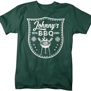 May include: Dark green t-shirt with a white graphic. The graphic is a shield shape with the words "Johnny's BBQ ESTD. 1983". A grill, flames, and utensils are also featured in the design. The shirt is made of a soft material.