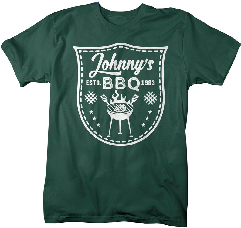 May include: Dark green t-shirt with a white graphic. The graphic is a shield shape with the words "Johnny's BBQ ESTD. 1983". A grill, flames, and utensils are also featured in the design. The shirt is made of a soft material.