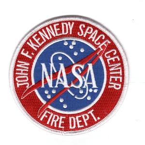 Puede incluir: Parche bordado con el logotipo de la NASA y el texto "JOHN F. KENNEDY SPACE CENTER FIRE DEPT.". El parche tiene un borde y texto rojos, con un centro azul que contiene el logotipo de la NASA y texto blanco.