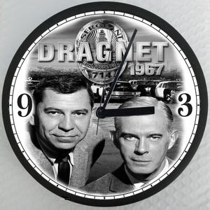 May include: Round black and white wall clock featuring the text "DRAGNET 1967" and images of two men. The clock face includes the numbers 9 and 3. The clock hands are black.