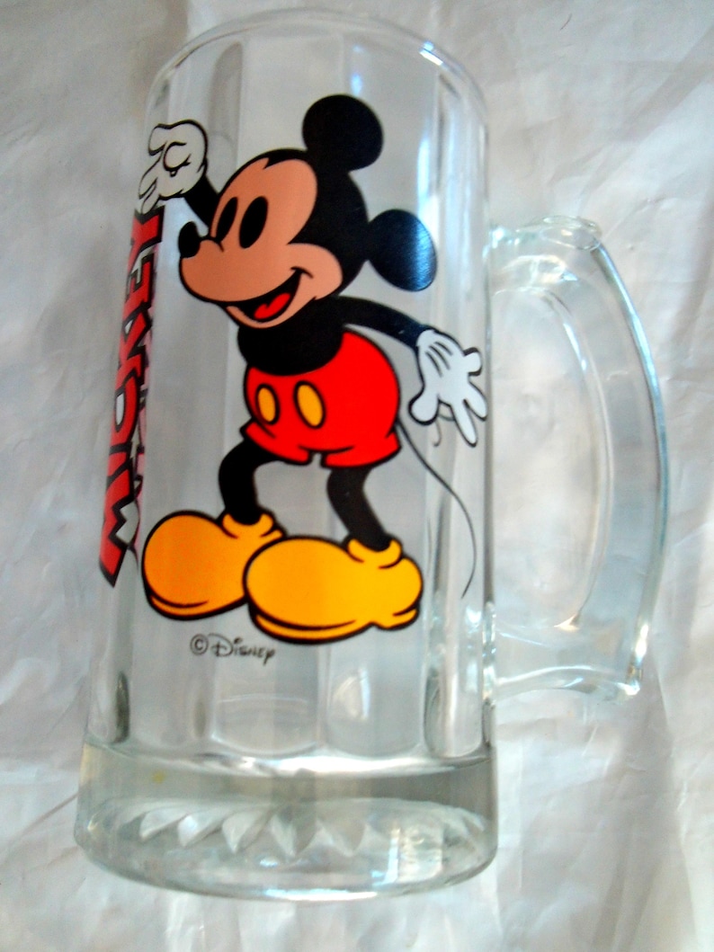 Puede incluir: Una jarra de cerveza de cristal transparente con un gr&aacute;fico colorido de Mickey Mouse con pantalones cortos rojos y zapatos amarillos. La palabra MICKEY est&aacute; impresa verticalmente en letras rojas junto al personaje. La base tiene un dise&ntilde;o facetado.