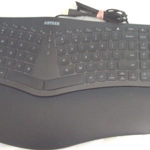 May include: A black Arteck ergonomic keyboard with a curved layout and an integrated wrist rest. The keyboard features a standard QWERTY key arrangement, a numeric keypad on the right side, and a black USB cable coiled at the top center.