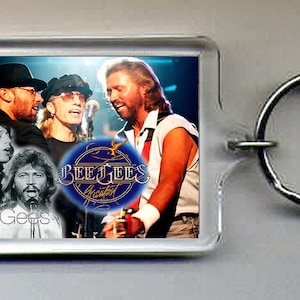May include: A clear plastic keychain with a photo of the Bee Gees band members. The photo shows the band members in a concert setting. The keychain has a silver metal ring.