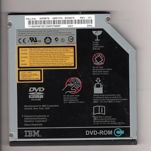 May include: An IBM DVD-ROM multi-player optical drive with a black and silver metal casing. The top surface features a white label with part numbers 92P6579 and 92P6578, a barcode, and various safety warning icons and text in multiple languages.