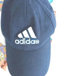 May include: Navy blue baseball cap with the Adidas logo in white. The cap has a curved brim and a structured crown. The Adidas logo features three white stripes above the brand name. This cap is a classic athletic accessory.