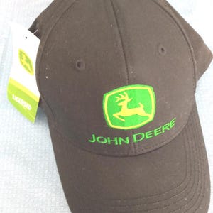 May include: A brown baseball cap featuring the John Deere logo in green and yellow. The logo includes a deer inside a green shield, with the words "JOHN DEERE" below. The cap has a curved brim and a button on top.