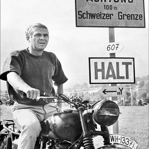 Può includere: Una fotografia in bianco e nero di un uomo su una motocicletta. Indossa una felpa e guida una motocicletta con una targa che recita "WH-1337". La motocicletta è ferma a un cartello che recita "ACHTUNG 100 m Schweizer Grenze" e "HALT".