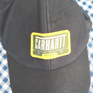 May include: Black baseball cap with a mesh back and a rectangular patch. The patch has the word "CARHARTT" in white letters on a green and yellow background. The patch also includes the text "QUALITY MADE - CRAFTED TO OUTLAST" and "GENUINE GOODS".