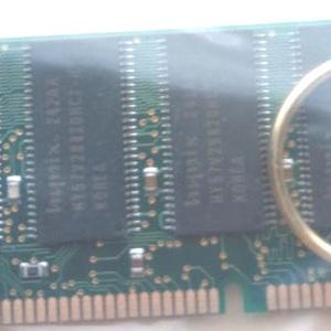 May include: A green circuit board with black rectangular chips and a gold-colored keyring attached. The board has small components and text. The keyring is circular and made of metal.