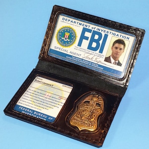 May include: A black leather FBI badge holder with a gold badge and an FBI identification card. The ID card features a photo, the text "Special Agent", and the FBI seal. The holder is open, revealing the contents.