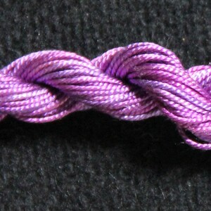 May include: A skein of Ebb Tide embroidery floss in shades of purple and lavender. The floss is tightly twisted, creating a rope-like appearance. The label on the right side of the image reads "Ebb Tide". The background is a dark, textured fabric.