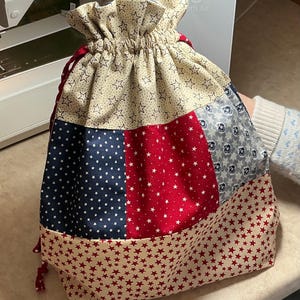 May include: A patchwork drawstring bag with a patriotic theme. The bag features panels of navy blue with white stars, red with white stars, and beige with red stars. The top is gathered with a drawstring closure.