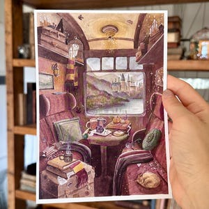 May include: A rectangular art print depicting the interior of a train carriage with plush red seats, a central table, and a large window showing a castle landscape. The scene includes a white owl in a cage, books, mugs, and a striped scarf.