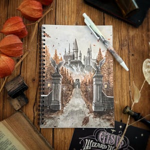 Peut inclure: Un carnet à spirales avec une peinture à l'aquarelle d'un château sur une colline, avec un chemin menant à celui-ci. L'image est entourée de fleurs orange séchées, d'un stylo et d'articles de papeterie sur une surface en bois.
