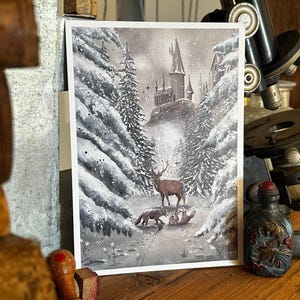 May include: A watercolor print depicting a snowy landscape with a castle in the distance. A deer and a fox are in the foreground. The artwork is framed in white and set against a wooden surface.