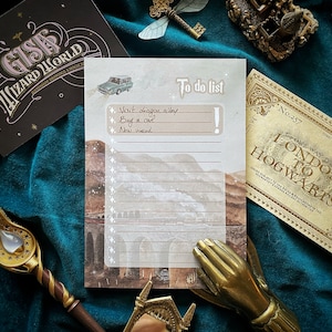 Può includere: Un blocco note con una lista di cose da fare con un'illustrazione ad acquerello di un ponte ferroviario e un'auto. Il blocco note contiene un elenco di cose da fare, tra cui "Visita il Vicolo Diagon", "Compra un gufo" e "Nuova bacchetta magica".