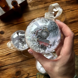 Puede incluir: Una bola de Navidad de cristal transparente con una escena de tren en su interior. El adorno tiene una cinta blanca para colgar. El tren es rojo y negro, sobre un fondo de bosque nevado. El adorno lo sostiene una persona.