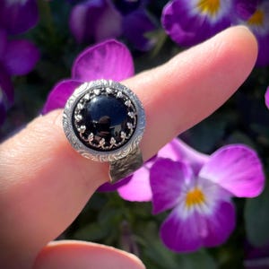 Op de afbeelding: Een zilveren ring met een grote, ronde, zwarte edelsteen. De ring heeft een sierlijke, gedetailleerde zetting met een bloemmotief. De achtergrond toont paarse bloemen.