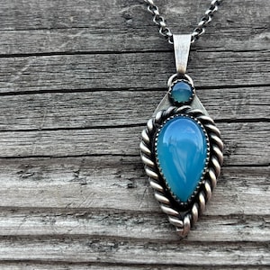 Op de afbeelding: Zilveren ketting met een lichtblauwe, druppelvormige hanger. De hanger heeft een gedraaid touwontwerp rond de randen en een kleinere blauwe steen erboven. De ketting is een fijne zilveren schakel.