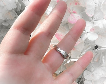 May include: A silver ring with a textured, irregular design. The ring is worn on a finger against a background of white flowers.