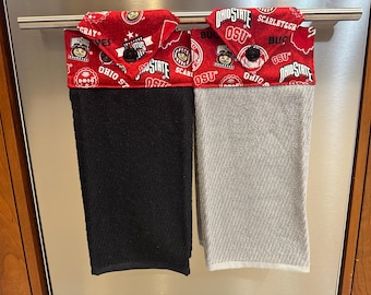 May include: Two kitchen towels hanging on a rack. The towel on the left is black with a red and white Ohio State University themed border. The towel on the right is gray with a red and white Ohio State University themed border.