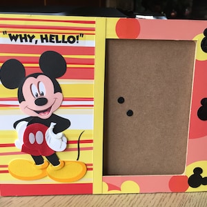 Könnte beinhalten: Ein gelber, roter und weißer Bilderrahmen mit einer Mickey-Maus-Figur auf der linken Seite. Der Rahmen trägt den Text "WHY, HELLO!" auf der linken Seite. Der Rahmen hat einen braunen Karton-Einsatz für ein Foto.