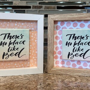 Puede incluir: Dos obras de arte enmarcadas con el texto "There's no place like bed". Un marco es blanco, el otro es de color madera. El fondo de cada pieza presenta un estampado floral o de lunares en tonos melocotón y rosa.