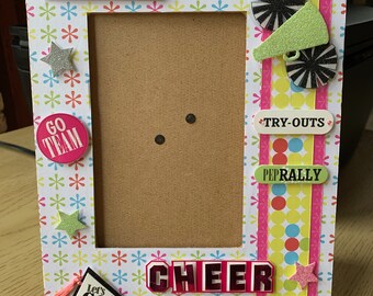 May include: A pink and white picture frame with a brown cardboard insert. The frame is decorated with colorful stars, pom-poms, and cheerleading-themed cutouts. The text "teamSPIRIT", "GO TEAM", "TRY-OUTS", "PEPRALLY", "Let's GO!" and "CHEER LEADER" is visible on the frame.