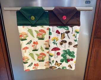 Op de afbeelding: Twee keukendoeken hangen aan een rek. De handdoek aan de linkerkant heeft een groene bovenkant en een witte onderkant met een paddenstoelmotief. De handdoek aan de rechterkant heeft een bruine bovenkant en een witte onderkant met een paddenstoel- en vlindermotief.