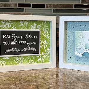 Puede incluir: Tres obras de arte enmarcadas con marcos blancos. Cada marco contiene un fondo decorativo y una pieza rectangular más pequeña con texto. El texto en las piezas dice "May God bless you and keep you" y "May the Lord bless & keep you."