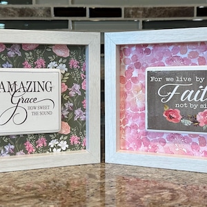 Puede incluir: Dos obras de arte enmarcadas con marcos blancos. Una presenta una cruz con detalles florales y el texto "Amazing Grace". La otra muestra el texto "For we live by Faith not by sight" con detalles florales. Ambos tienen fondos florales.