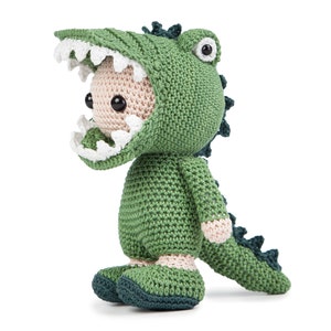 May include: A green crocheted alligator costume with a white mouth and black eyes. The costume is worn by a small crocheted figure with a light brown body.
