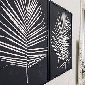 May include: Two black framed canvas prints with white palm leaf designs. The prints are hung on a white wall.