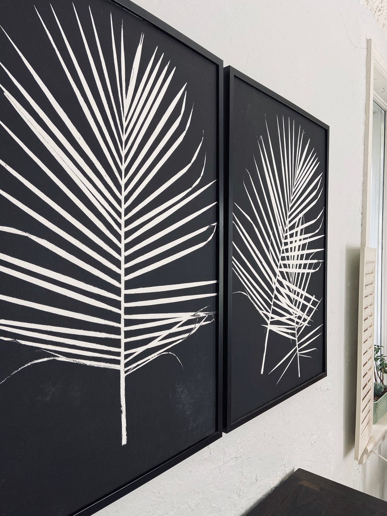 May include: Two black framed canvas prints with white palm leaf designs. The prints are hung on a white wall.