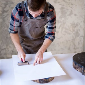 Op de afbeelding: Een persoon met een bruine leren schort en een geruit overhemd gebruikt een roller om een ​​vel wit papier glad te strijken op een houten oppervlak.