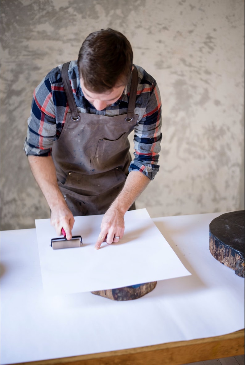 Op de afbeelding: Een persoon met een bruine leren schort en een geruit overhemd gebruikt een roller om een ​​vel wit papier glad te strijken op een houten oppervlak.