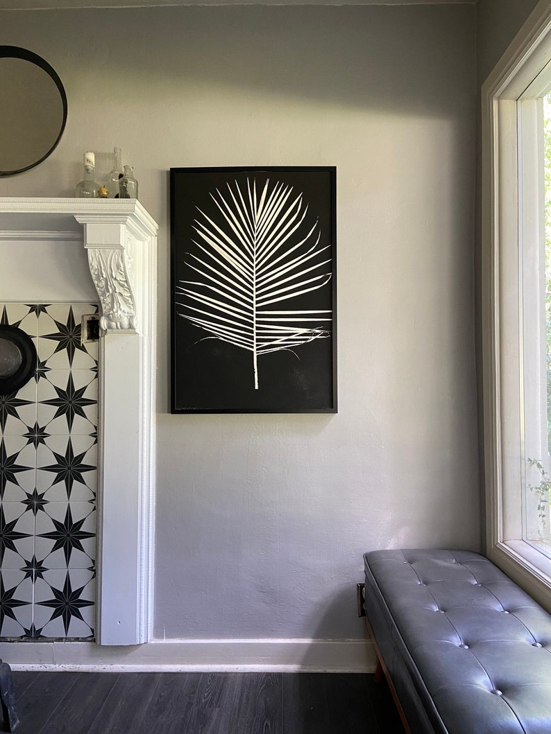 May include: A black framed print of a white palm leaf on a black background hanging on a light gray wall above a black leather bench.