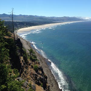May include: A panoramic view of a rocky coastline with a sandy beach and turquoise ocean water. The coastline is covered in green trees and shrubs. The sky is a clear blue.