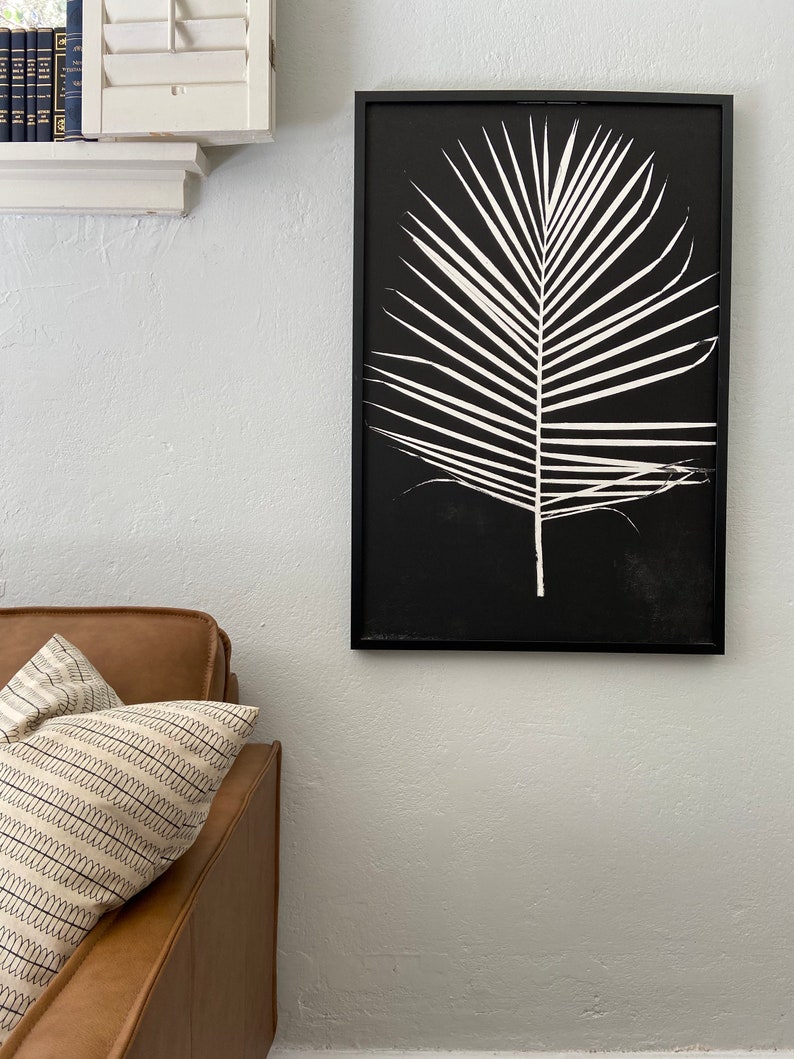 May include: A black and white framed print of a palm leaf on a black background. The print is hanging on a white wall.