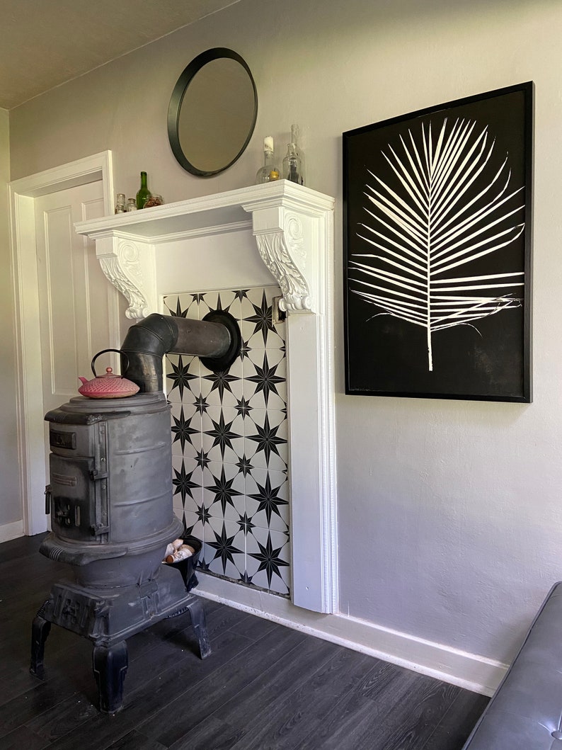 May include: A black wood-burning stove with a red teapot on top, set against a white and black tiled wall. A black framed print of a white palm leaf hangs on the wall above the stove.
