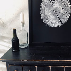 May include: A black dresser with multiple drawers, each with a wooden knob. On top of the dresser is a framed black and white print of a tree trunk cross-section. There are also two glass bottles and a wine bottle on the dresser.